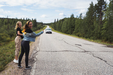 Finnland, Lappland, zwei glückliche Frauen, die per Anhalter auf einer Landstraße fahren - KKAF02081