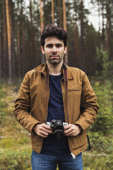 Finnland, Lappland, Porträt eines jungen Mannes mit Kamera in ländlicher Landschaft - KKAF02091
