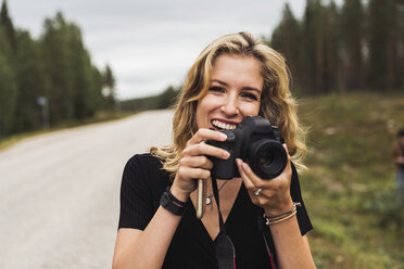 Finnland, Lappland, Porträt einer glücklichen jungen Frau mit Kamera an einer Landstraße - KKAF02094