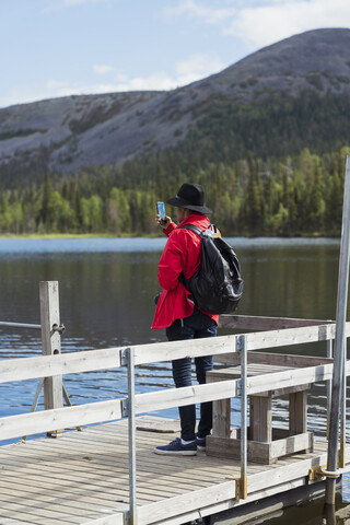 Finnland, Lappland, Mann fotografiert mit Handy am Seeufer, lizenzfreies Stockfoto