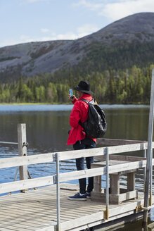 Finnland, Lappland, Mann fotografiert mit Handy am Seeufer - KKAF02108