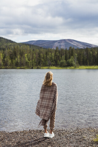 Finnland, Lappland, Frau in eine Decke gehüllt am Seeufer stehend, lizenzfreies Stockfoto