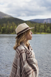 Finland, Lapland, woman wearing a hat wrapped in a blanket standing at the lakeside - KKAF02112