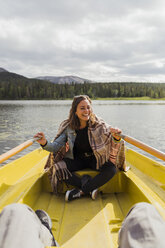 Finland, Lapland, laughing woman wearing a blanket in a rowing boat on a lake - KKAF02127