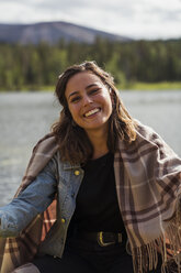 Finland, Lapland, portrait of smiling young woman wearing a blanket in a boat on a lake - KKAF02130