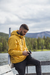 Finnland, Lappland, junger Mann mit einer Kamera auf einem Steg an einem See - KKAF02135