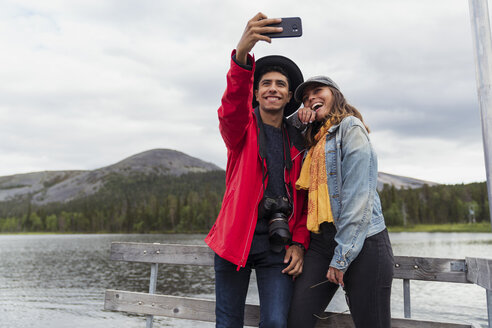 Finnland, Lappland, glückliches Paar auf einem Steg an einem See, das ein Selfie macht - KKAF02147