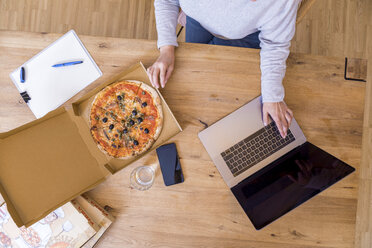 Frau im Büro zu Hause, die einen Laptop benutzt, während sie eine Pizza isst, Ansicht von oben - FMKF05305