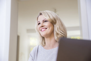 Porträt einer lächelnden blonden Frau mit Laptop zu Hause - FMKF05307