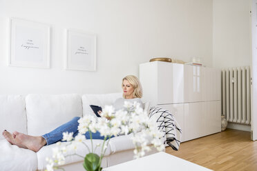 Blonde reife Frau sitzt auf der Couch im Wohnzimmer und benutzt ein digitales Tablet - FMKF05311