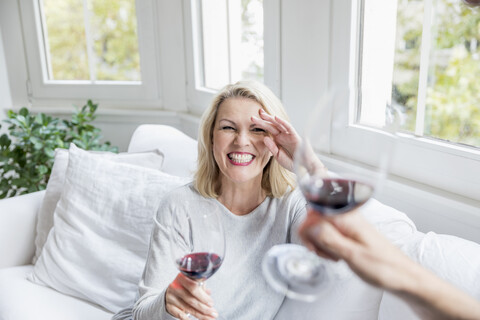 Porträt einer lachenden blonden reifen Frau, die zu Hause mit Rotwein anstößt, lizenzfreies Stockfoto