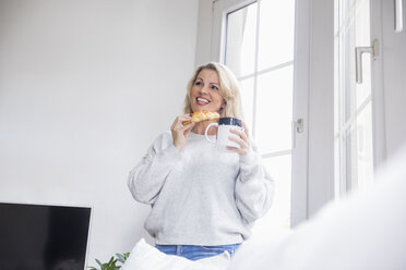 Porträt einer lächelnden blonden reifen Frau mit Croissant und Kaffeetasse zu Hause - FMKF05321