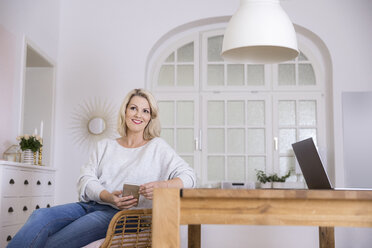 Portrait of smiling blond mature woman with smartphone and laptop at home - FMKF05322