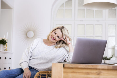 Porträt einer lächelnden blonden reifen Frau mit Laptop zu Hause - FMKF05325