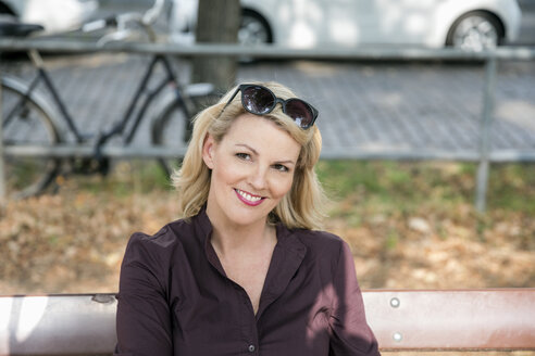 Porträt einer lächelnden blonden reifen Frau, die auf einer Bank im Freien sitzt - FMKF05331