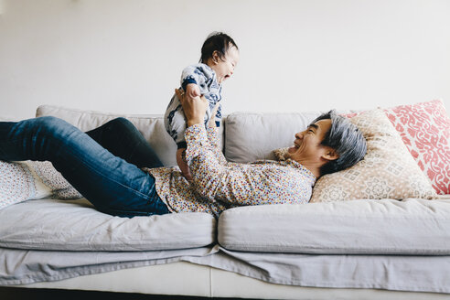 Glücklicher Großvater spielt mit seinem Enkel, während er zu Hause auf dem Sofa liegt - CAVF48964