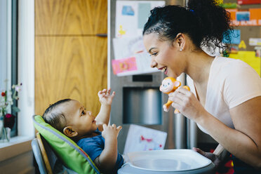 Playful mother playing with son sitting on high chair in kitchen at home - CAVF48966