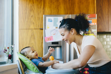 Mother playing with son sitting on high chair in kitchen - CAVF48967