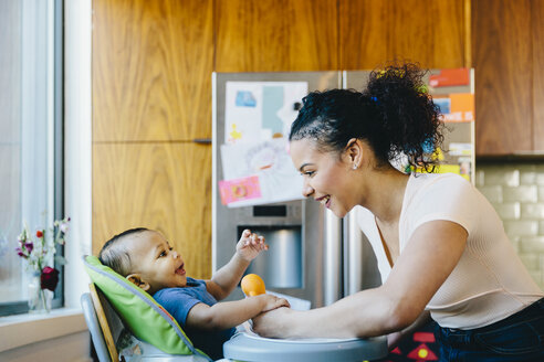 Mother playing with son sitting on high chair in kitchen - CAVF48967