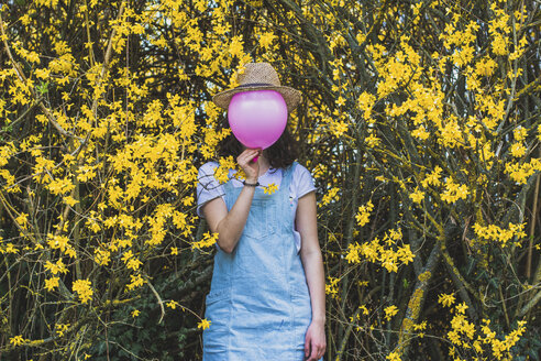 Playful young woman holding pink helium balloon with hat against face while standing by yellow blossoms at park - CAVF49017