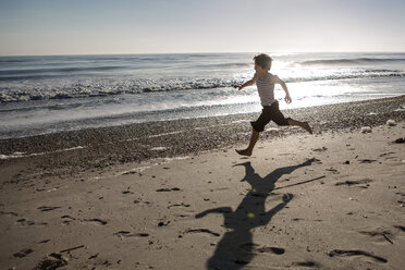 Voller Länge des spielerischen Jungen läuft am Strand bei Sonnenuntergang - CAVF49032