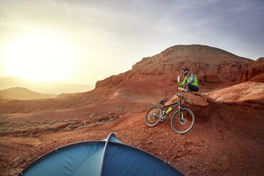 Wanderer trinkt Kaffee und sitzt mit seinem Mountainbike auf einem Felsen in der Wüste - CAVF49087