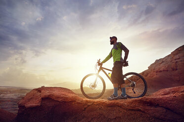 Full length of hiker with mountain bike standing on rocks against sky at desert during sunset - CAVF49092