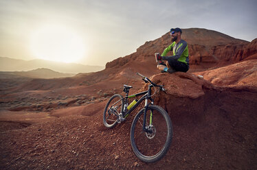 Full length of hiker by mountain bike sitting on rocks at desert during sunset - CAVF49093