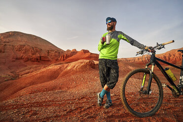 Wanderer hält ein Getränk in der Hand, während er mit seinem Mountainbike in der Wüste bei Sonnenuntergang steht - CAVF49094