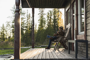 Junger Mann sitzt auf der Veranda eines Holzhauses und spielt auf der Ukulele - KKAF02174