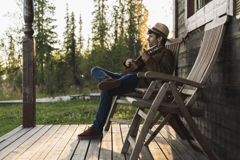 Junger Mann sitzt auf der Veranda eines Holzhauses und spielt auf der Ukulele - KKAF02175