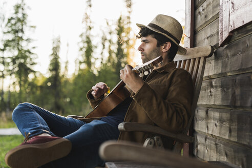 Junger Mann sitzt auf der Veranda eines Holzhauses und spielt auf der Ukulele - KKAF02176