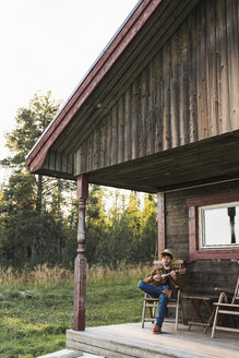 Junger Mann sitzt auf der Veranda eines Holzhauses und spielt auf der Ukulele - KKAF02178