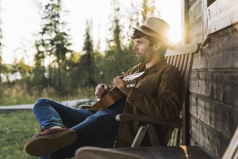 Young man sitting on veranda of a wood house, playing the ukulele - KKAF02180