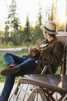 Junger Mann sitzt auf der Veranda eines Holzhauses und spielt auf der Ukulele - KKAF02181