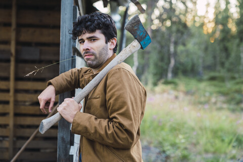 Junger Mann hält eine Axt und lehnt sich an ein Holzhaus, lizenzfreies Stockfoto