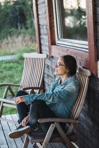 Glückliche junge Frau sitzt auf der Veranda eines Holzhauses, lizenzfreies Stockfoto