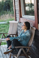 Happy young woman sitting on veranda of a wooden house - KKAF02225