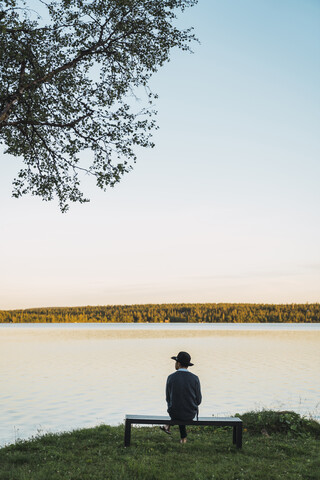 Junger Mann sitzt auf einer Bank und schaut auf den See, lizenzfreies Stockfoto