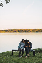 Junges Paar sitzt auf einer Bank in der Nähe eines Sees und prüft Bilder auf einer Kamera - KKAF02231