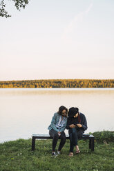 Young couple sitting on a bench near lake, checking pictures on a camera - KKAF02231