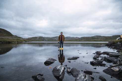 Young man standing ankle deep in water, holding camera - KKAF02308