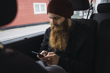 Bärtiger junger Mann sitzt auf dem Rücksitz eines Autos und überprüft sein Handy - KKAF02313