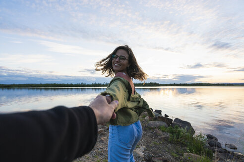 Finland, Lapland, happy young woman holding man's hand at the lakeside at twilight - KKAF02321