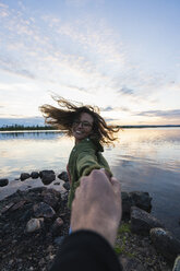 Finland, Lapland, happy young woman holding man's hand at the lakeside at twilight - KKAF02322