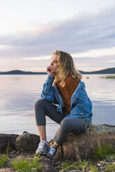 Finland, Lapland, young woman sitting on a rock at the lakeside - KKAF02326