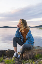 Finnland, Lappland, junge Frau sitzt auf einem Felsen am Seeufer - KKAF02326
