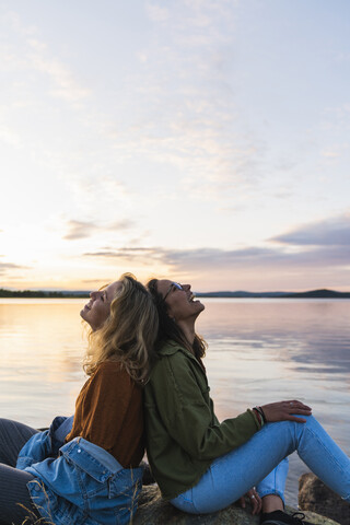 Finnland, Lappland, zwei glückliche junge Frauen sitzen in der Dämmerung Rücken an Rücken am Seeufer, lizenzfreies Stockfoto