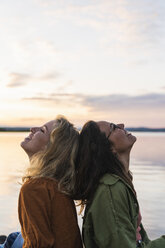 Finnland, Lappland, zwei glückliche junge Frauen Rücken an Rücken am Seeufer in der Dämmerung - KKAF02329