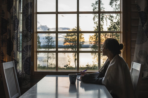 Finland, Lapland, young woman sitting at the window looking at a lake - KKAF02342